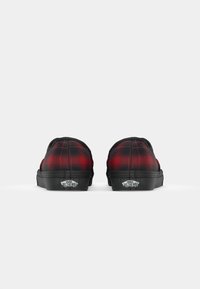 Zapatillas slip-on de cuadros rojos y negros con suela de goma negra y etiqueta de marca en el talón. La parte superior presenta un acabado de tela texturizada.