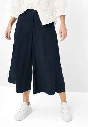 Kvinde iført løse marineblå culottes, hvid blomsterbluse og hvide sneakers, stående mod en hvid baggrund.