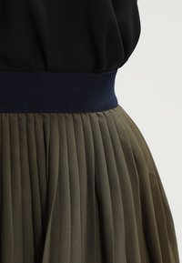 Jupe plissée vert olive avec une large ceinture bleu marine. Le tissu a une texture douce avec un léger éclat et un design fluide.