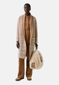 Beige Strickjacke mit strukturiertem Muster, langen Ärmeln und zwei Taschen, über einem braunen Hemd und passenden Hosen getragen. Enthält eine flauschige cremefarbene Tasche.