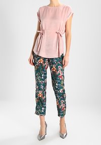 Top corto rosa chiaro a maniche corte con dettagli regolabili a fiocco, abbinato a pantaloni con stampa floreale verde e scarpe con tacco alto argentate.