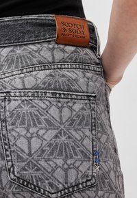 Gray patterned denim jeans featuring a brown leather label with "SCOTCH & SODA AMSTERDAM." Blue and gray stitching accents on the pocket.