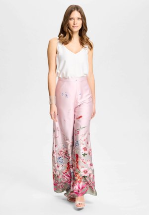 Vrouw met golvend haar, die een witte mouwloze top draagt, roze wijde bloemenbroek, beige open tenen hakken en een zilveren armband, staand.