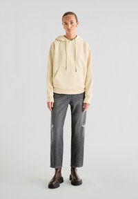 Lichtbeige hoodie met een voorzak en koordjes, gecombineerd met versleten zwarte spijkerbroek en stevige zwarte laarzen.