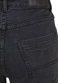 Jeans de denim negro con una vista cercana del área del bolsillo trasero, con una textura suave y sutiles detalles de costura.
