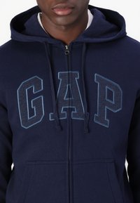 Sweat à capuche zippé bleu marine avec un grand logo "GAP" en bleu plus clair, capuche avec cordon de serrage et poches avant. Matière douce avec coutures plates.