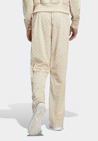 Béžové jogger kalhoty s jemným vzorem listů, elastickým pasem a manžetami, se skládacími kapsami a bílou podrážkou. Hladká textura.