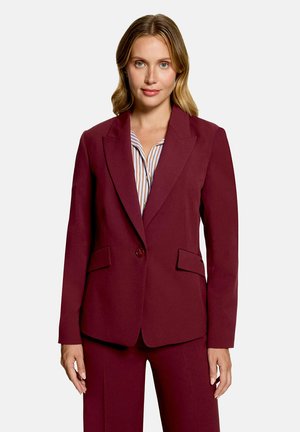 Femme portant un blazer bordeaux ajusté avec un bouton fermé sur une chemise à rayures, associé à un pantalon bordeaux assorti.