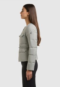 Femme aux longs cheveux bruns portant une doudoune ceinturée gris clair et un pantalon noir, debout de profil devant un fond uni.