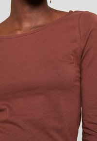 Top lungo marrone a maniche lunghe realizzato in cotone morbido. Presenta un collo rotondo e una silhouette aderente con dettagli minimi.