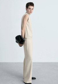 Bež bezrukavni pulover s visokim ovratnikom uparen s odgovarajućim širom hlačama, držan crnom clutch torbicom. Glatka tekstura, minimalisti�čki dizajn.