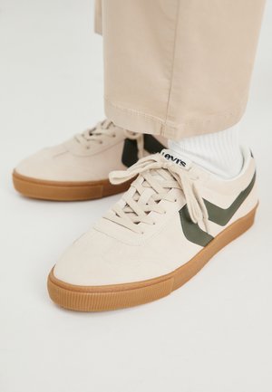 Beige Wildledersneaker mit grünen Lederakzenten, Schnürdesign und Gummisohle. Weiße Socke über dem Knöchel sichtbar, kombiniert mit beigen Hosen.