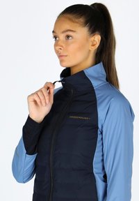 Navy and light blue jacket with a high collar, zip closure, and logo on the front. Textured material with contrasting sleeve colors.