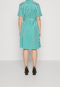 Teal- ja valkoisraidallinen lyhythihainen mekko, jossa on tiukka vyötärö ja levenevä hameosa. Takana näkyy vyöllinen muotoilu ja sileä pinta.