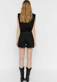 Femme aux longs cheveux blonds portant un haut noir sans manches, un short noir retroussé et des bottes noires mi-mollet, debout de dos.