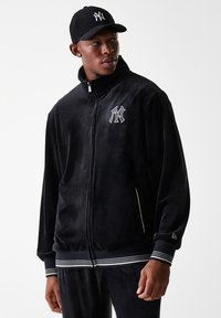 Veste en velours noir avec fermeture éclair sur le devant, col montant et logo NYC brodé en blanc. Comprend des poignets et une taille rayés pour le contraste. Casquette également représentée.