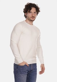 Maglione a maniche lunghe di colore crema con scollatura a girocollo, realizzato in un tessuto a maglia morbida, abbinato a jeans di colore blu scuro.