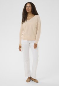 Pull en maille beige clair avec col en V, assorti à un pantalon blanc à jambes droites et des chaussures en toile tressée couleur bronze. Design simple et texturé.