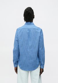 Mężczyzna o ciemnej karnacji z dredami, ubrany w niebieską, pionowo paskowaną koszulę z długim rękawem oraz jasne spodnie, stojący tyłem.