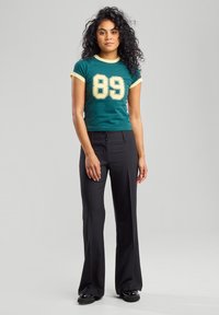 Vihreä lyhythihainen t-paita, jossa on keltainen reunus ja rohkea "89" -graafinen printti, yhdistettynä mustiin leveneviin housuihin, joissa on napitettu etuosadon.