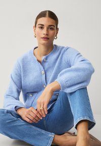 Lichtblauwe gebreide cardigan met knoopsluiting, ronde halslijn en ballonmouwen, gecombineerd met lichtblauwe rechte jeans.