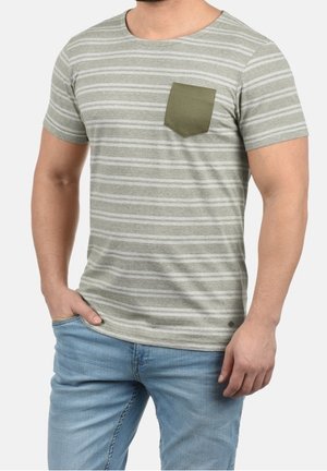 Mand iført en kortærmet, grå stribet t-shirt med lomme på brystet i olivengrøn og lyseblå jeans, stående med den ene hånd i lommen.