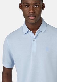Polo de color azul claro fabricado en tejido texturizado, con una tapeta de dos botones y un acento de logo en el pecho izquierdo. Mangas cortas.