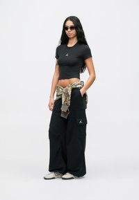 Γυναίκα με μαύρο κοντό μπλουζάκι, φαρδιά μαύρα cargo παντελόνια, αλεξίσφαιρο μπουφάν σε παραλλαγή δεμένο στη μέση, λευκά sneakers και γυαλιά ηλίου, στέκεται μπροστά σε λευκό φόντο.