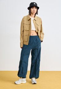 Κάι πιτζάμα επάνω πανωφόρι με τέσσερις τσέπες, κρεμ κοντό τοπ, τυρκουάζ χαλαρά παντελόνια, μαύρο καπέλο κα bucket hat και ανακλαστικά αθλητικά παπούτσια.