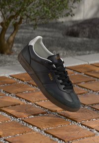 Sneakers din piele neagră cu accente din suede verde, interior alb și șireturi negre. Prezintă logo-ul auriu "BOSS" și o talpă din cauciuc texturat.
