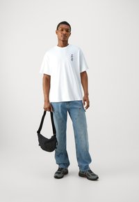 White short-sleeve t-shirt with black graphic on the chest, paired with light blue baggy jeans and black crossbody bag, worn with gray sneakers.