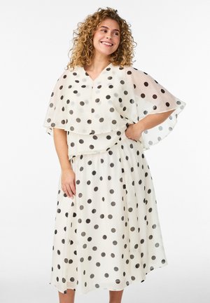 Usmievavá žena s kučeravými vlasmi, oblečená v bielych šatách s čiernymi bodkami, ktoré majú plášťový vrch a prúdiacu sukňu.
