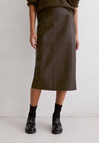 Brauner Leder-Midirock mit geradem Schnitt, ausgestattet mit vorderen Einsätzen und Seitentaschen. Kombiniert mit schwarzen Socken und Slippern.