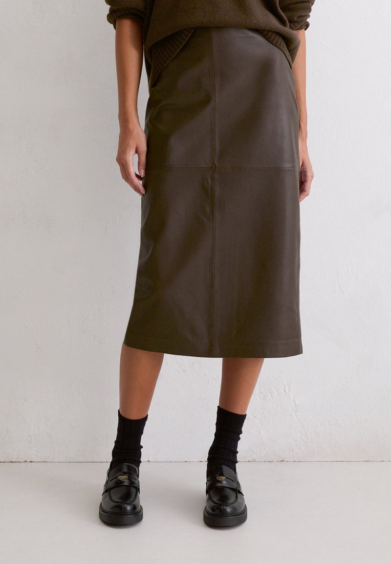 Brauner Leder-Midirock mit geradem Schnitt, ausgestattet mit vorderen Einsätzen und Seitentaschen. Kombiniert mit schwarzen Socken und Slippern.
