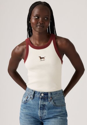 Jeune femme aux cheveux tressés porte un débardeur blanc avec un liseré rouge et un petit motif chien, assorti à un jean en denim bleu.