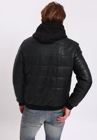 Černá kožená bunda s vypolstrovaným designem a žebrovanými manžetami, která má černou mikinu připevněnou k límci a modré džíny.