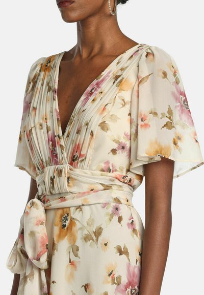 Nő krémszínű virágos ruhában, pliszírozott V-nyakkal, fodros ujjakkal és széles derékpánttal, amely oldalt masniba van kötve.