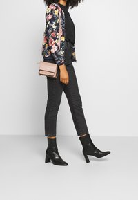 Veste bomber florale avec un fond bleu marine, haut noir, jeans slim noirs et bottines à talons noires. Tenant un petit sac bandoulière rose.