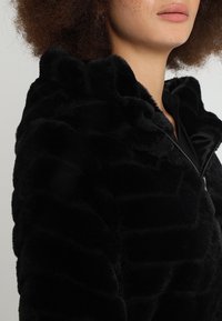 Černá umělá kožešinová bunda s texturovaným vzorem, zipovým zapínáním a kapucí. Tkanina vypadá jemně a nadýchaně, což zdůrazňuje rozměr designu.