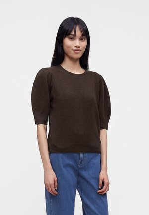 Jeune femme aux cheveux noirs lisses porte un pull marron à manches bouffantes et un jean bleu, debout devant un fond clair uni.