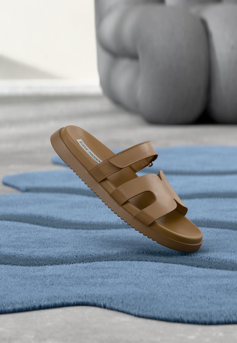 Sandale marron en cuir à enfiler avec un design découpé et une bride ajustable, posée sur un tapis bleu texturé avec un arrière-plan gris flou.