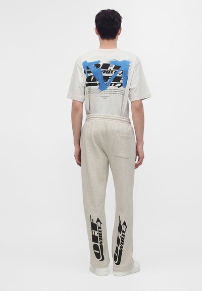 Homme de dos portant un t-shirt graphique gris clair et un pantalon de jogging assorti avec des logos Off-White noirs sur les mollets, sur un fond uni.
