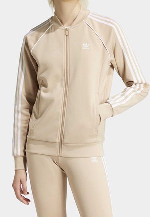 Beige Adidas trainingsjack en legging set met witte strepen, rits aan de voorkant en logo op borst en dij. Handen in de zakken.