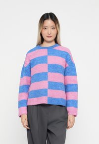 Ženska nosi pleten jumper z debelo pletenjem, ki ima izmenične roza in modre široke vodoravne proge, v kombinaciji s širokimi temno sivo hlačami.