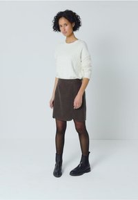 Witte kabelgebreide trui, bruine A-lijn rok van textuurstof, zwarte enkellaarsjes en zwarte panty tegen een neutrale achtergrond.