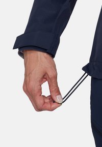 Hand die de trekkoordzoom van een marineblauw jasje verstelt, met de mouwboord met klittenbandsluiting zichtbaar tegen een eenvoudige achtergrond.