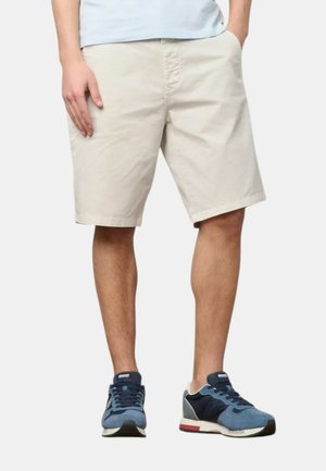 Uomo che indossa pantaloncini beige fino al ginocchio, sneakers azzurri con dettagli blu scuro, in piedi con una mano in tasca.