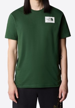 Osoba w ciemnozielonym t-shircie The North Face z małą białą łatką z logo na lewej piersi, w połączeniu z czarnymi spodniami.