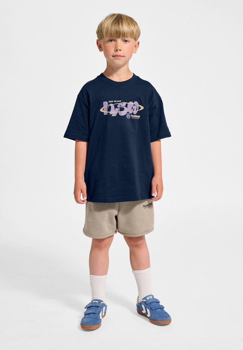 Blonder Junge steht und trägt ein marineblaues T-Shirt mit Grafik, beige Shorts, weiße Socken und blaue Sneakers vor weißem Hintergrund.