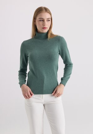 Femme aux longs cheveux blonds portant un pull à col roulé vert et un pantalon blanc, regardant légèrement sur le côté sur un fond uni.